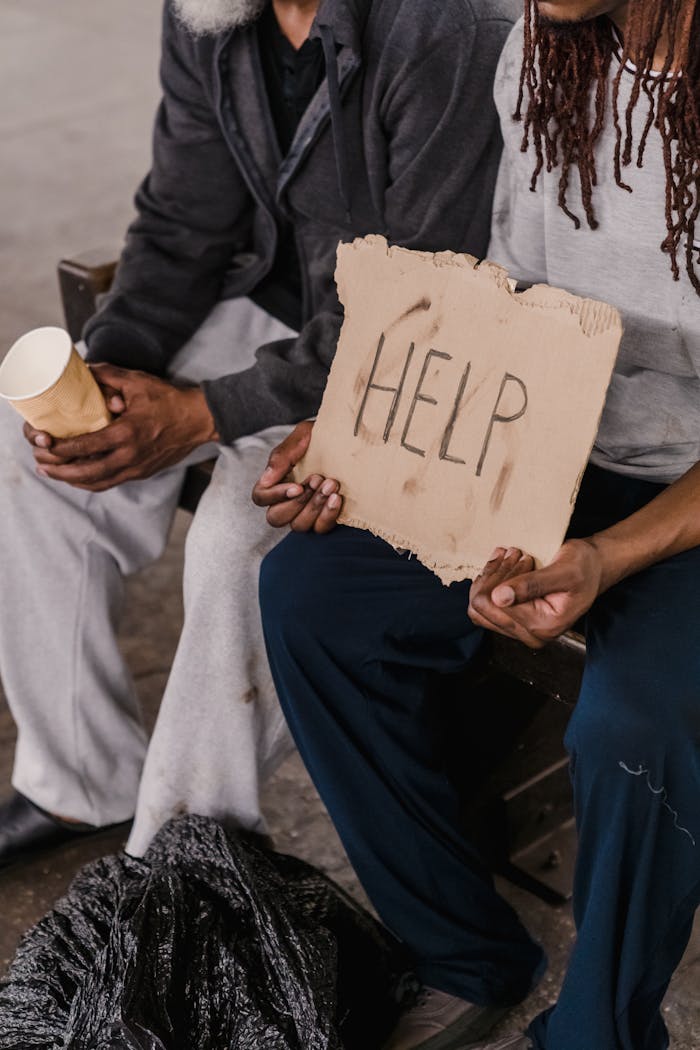 Two homeless individuals hold a cardboard sign with 'HELP' written, depicting a plea for assistance.
